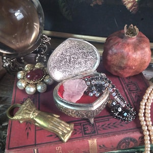 May include: A heart-shaped silver jewelry box with an open lid, revealing a pink crystal and black beaded jewelry. A gold hand-shaped bottle opener and a red and gold brooch are also visible. A pomegranate and a strand of pearls are also in the image.