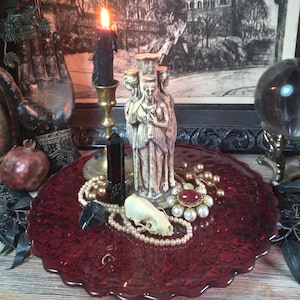 May include: A gothic-themed display featuring a lit black candle in a brass holder, a statue of three figures, and a red glass plate. The plate holds a skull, pearls, and a red brooch. A framed black and white print of a cathedral is in the background.