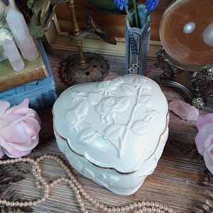 May include: A heart-shaped, white ceramic trinket box with a gold trim and embossed floral design. The box is displayed with a pearl necklace, pink roses, and a small vase of blue flowers. A crystal ball and other decorative items are in the background.