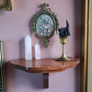 May include: A semi-circular wooden wall shelf holding two clear quartz crystal points and a brass candle holder with a black wax sculpture. Above the shelf hangs an oval-shaped ornate gold-toned metal frame containing a floral print.