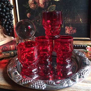 May include: Four ruby red, faceted drinking glasses sit on a silver serving tray. The glasses have a textured, geometric design. A clear glass sphere and a framed floral painting are in the background. The tray is on a wooden surface.