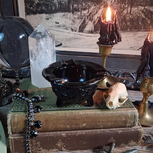 May include: A dark brown, three-legged glass ashtray with a starburst pattern on its exterior. The ashtray features a wide, circular rim with integrated notches for resting items, and it is positioned centrally on top of two stacked vintage books.