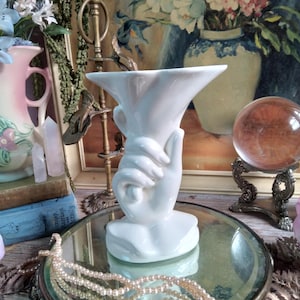 May include: A white ceramic vase in the shape of a hand holding the vase. The vase is on a mirrored surface with a pearl necklace. Other items in the background include a pink vase, books, and a painting of flowers.