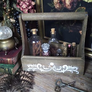 May include: A rustic wooden caddy with a handle, holding several glass bottles and vials. The caddy is decorated with a white floral design. The bottles contain various spices and herbs. A vintage aesthetic is present with old keys and antique books.