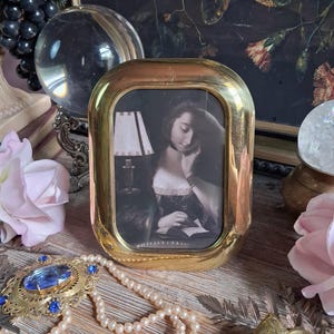 May include: An oval-shaped polished brass picture frame containing a black and white print of a person reading by a lamp. The frame has a smooth, reflective metallic surface and a rounded rectangular aperture, showcasing a classic portrait style.