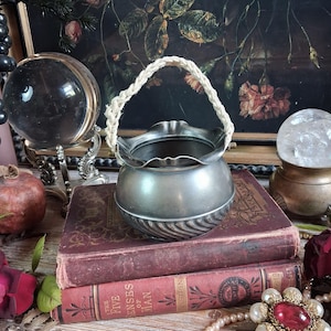 May include: A round, silver-toned metal vessel with a ruffled rim and a braided cream-colored cord handle. The base of the vessel features a decorative diagonal ribbed pattern. The object is resting on top of two stacked vintage books.