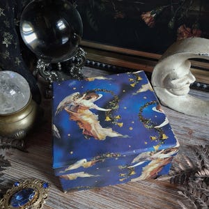 May include: A square box featuring a deep blue night sky background with illustrations of winged figures in flowing robes, golden bells, and yellow stars. The box lid and sides are decorated with this celestial pattern, showing multiple angels.