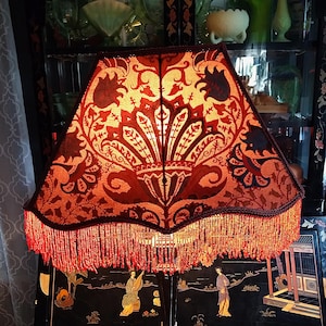 May include: A lampshade with a red and gold floral design and a beaded fringe. The lampshade is the central element, emitting a warm light. The background features a black cabinet with glass shelves and various decorative objects.