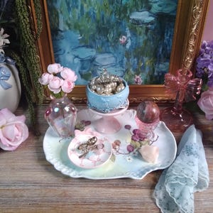 May include: A still life arrangement featuring a framed painting, a small blue cake stand with pearls, a decorative tray with small dishes, a pink glass vase, and a decorative pink cross. The color palette includes pink, blue, and gold.