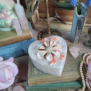 May include: A heart-shaped, white trinket box with a seashell and coral-colored ribbon design sits atop vintage books. The image also features a blue vase with blue flowers, quartz crystals, and a pearl necklace, creating a vintage aesthetic.