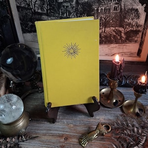 May include: A bright yellow hardcover book featuring a centered black line art illustration of a sun with a human face. The book is displayed upright on a dark brown stand, showcasing a clean, solid-colored front cover with a minimalist sun icon.