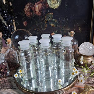 May include: A collection of six clear glass apothecary bottles with white plastic stoppers, arranged on a round mirrored tray. The bottles are cylindrical with a narrow neck and a wide base, reflecting light against the reflective surface.
