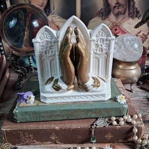 May include: A white ceramic figurine depicting two hands pressed together in prayer, featuring gold-toned accents on the hands and cuffs. The hands are positioned in front of a white arched architectural backdrop with embossed floral details.