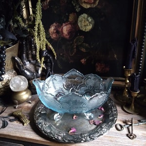 May include: A light blue glass bowl with a scalloped edge and intricate floral design sits on a silver tray with a floral pattern. The bowl has three feet and is surrounded by rose petals.
