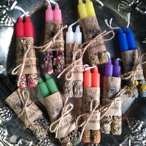 May include: A collection of colorful spell candles, each with two wicks, wrapped in brown paper with an eye design and tied with twine. The candles are various colors, including red, pink, yellow, white, blue, green, orange, purple, and black.