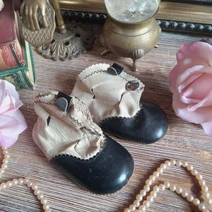 May include: A pair of vintage baby shoes with black soles and cream-colored uppers. The shoes have a scalloped edge and small black straps. The image also includes a crystal ball, a pearl necklace, and a pink rose.