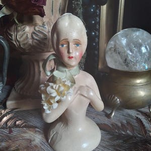 May include: A vintage porcelain doll bust with a green ribbon around its neck and a bouquet of white flowers in its hand. The doll has painted blue eyes, red lips, and a white face.