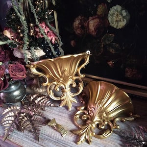 May include: Two gold-coloured wall shelves with ornate scrollwork and a shell-like design. The shelves are made of a lightweight material and have a distressed finish.
