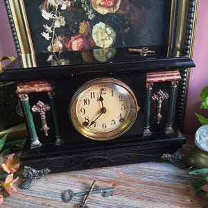 May include: A black antique mantel clock with a round clock face and gold-colored trim. The clock has decorative columns and ornate details. Two keys are placed in front of the clock. The clock is set against a backdrop of a framed floral painting.
