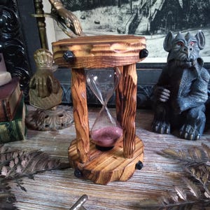 May include: A wooden hourglass with a clear glass center filled with pink sand. The hourglass is supported by a wooden frame with a square top and bottom. The wood has a natural, light brown finish. Black accents are on the top and bottom.