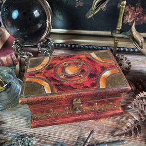 May include: A small, antique wooden box with a red and orange patterned top and brass accents. The box is rectangular with a hinged lid and a small lock. Two antique keys are in front of the box. A crystal ball and other vintage items are in the background.