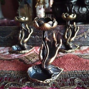 May include: Bronze-toned candle holders in the shape of hands holding lotus flowers. The holders have a dark, antique finish and sit on a decorative base. The background features a red and gold patterned fabric.