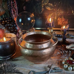 May include: A hammered copper cauldron with a rounded body and a curved metal handle. The pot features a wide opening and a textured metallic surface. It is positioned in front of a dark floral painting, a crystal ball, and two lit candles.
