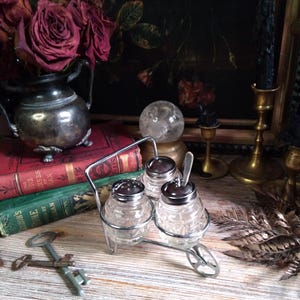 May include: A vintage silver-tone condiment set with three glass jars and a small spoon. The jars are in a wheeled cart design and have silver-tone lids. The set is on a wooden surface with keys and a stack of books.