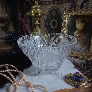 May include: A clear, cut-glass bowl featuring a scalloped rim and intricate floral etchings on the exterior. The bowl is supported by three small, curved glass feet and displays a faceted pattern that catches light across its transparent surface.