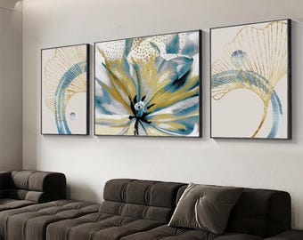 May include: Three framed abstract art pieces. The artwork features floral and botanical designs in shades of blue, gold, and white. The frames are black. The art is displayed above a brown sectional sofa.