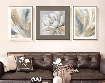 May include: A triptych of framed abstract art pieces. The central piece features a large white flower painting, flanked by two abstract paintings with blue, brown, and white brushstrokes. The artwork hangs above a brown leather sofa.