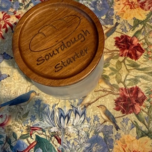May include: A wooden lid with the words "Sourdough Starter" engraved on it, covering a glass jar. The lid features a bread loaf design. The jar sits on a floral patterned fabric with birds and flowers.