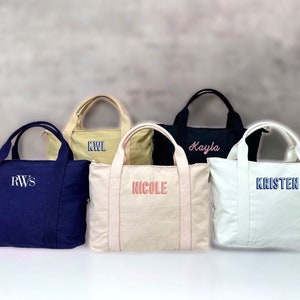 May include: Five personalized tote bags in various colors: navy blue, beige, black, cream, and off-white. Each bag features embroidered text in different colors, including initials and names. The bags have double handles and a rectangular shape.