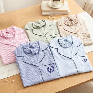 May include: Five folded button-down pajama shirts with matching scrunchies in pink, green, tan, and two shades of blue stripes. Each shirt features a notched collar, a chest pocket, and embroidered initials or names including SVZ, IGB, A, and KER.
