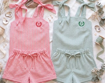 May include: Two seersucker short sets in red and green stripes. Each set includes a tank top with tie-shoulder straps and matching shorts with a drawstring waist. The red top features a red embroidered letter T, and the green top features a green embroidered letter P. Two matching fabric scrunchies are included.