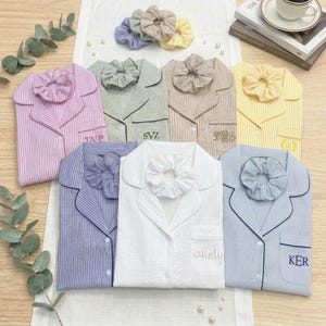 May include: An assortment of striped pajama sets in various colors, including pink, green, beige, yellow, blue, and white, each paired with a matching scrunchie. Some pajamas have embroidered initials. The sets are arranged on a white surface.