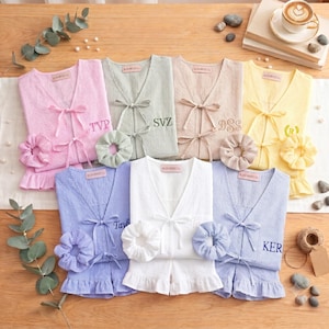 Può includere: Sette set di pigiami coordinati in rosa, verde, beige, giallo, blu e bianco, ognuno composto da un top con scollo a V e laccetti frontali, pantaloncini con volant e un elastico per capelli abbinato. Ogni top è ricamato con iniziali o nomi come TVP, SVZ, DSS e KER.
