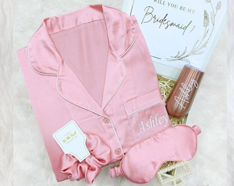 May include: A pink satin pajama top with white piping, a matching pink satin sleep mask, a pink satin scrunchie, a small white card with gold lettering that says "I don't do the bride without you", and a rose gold tumbler with the name "Ashley" engraved on it. The items are arranged in a gift box with a white lid that says "Will you be my Bridesmaid?" in gold lettering.