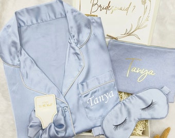 May include: A light blue satin pajama set with a sleep mask, a small pouch, and a hair scrunchie. The set is personalized with the name "Tanya" and the pouch has the text "Will you be my bridesmaid?" on a card.