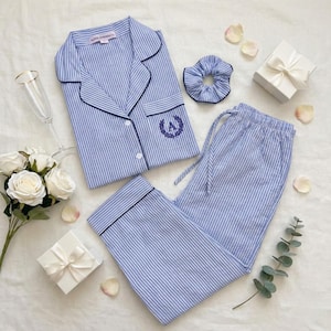 May include: Blue and white striped pajama set with a matching scrunchie and a gift box. The pajama top has a navy trim and an embroidered initial. The set includes a button-down shirt and drawstring pants. A champagne flute and flowers are also in the image.