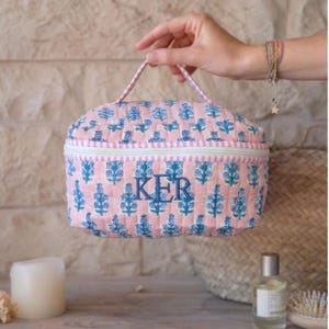 May include: A pink quilted cosmetic bag with a blue floral pattern and the embroidered initials "KER". The bag has a white zipper, a striped trim, and a matching handle. The bag is held by a person.