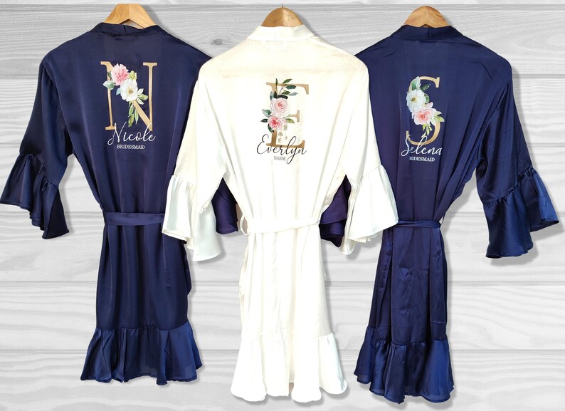 May include: Three satin robes with ruffled sleeves and hems. The robes are navy blue, white, and navy blue. Each robe has a large gold initial with floral accents and the name "Nicole Bridesmaid", "Evelyn Bridesmaid", and "Selena Bridesmaid" printed on the back.