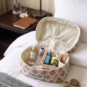 May include: Open floral-patterned cosmetic bag with various beauty products inside. The bag is cream-colored with pink flower designs and the initials "MVR" embroidered on the front. The bag is on a white bed with a nightstand in the background.
