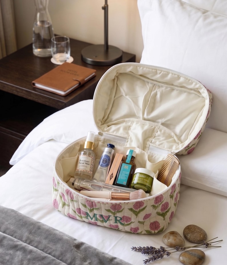 May include: Open floral-patterned cosmetic bag with various beauty products inside. The bag is cream-colored with pink flower designs and the initials "MVR" embroidered on the front. The bag is on a white bed with a nightstand in the background.