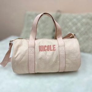 May include: A light pink canvas duffel bag with a gold zipper and two pink straps. The bag has the name "NICOLE" embroidered on the front in pink and red letters.