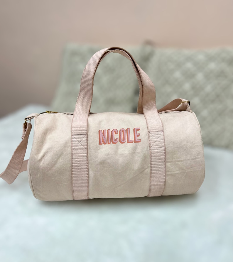 May include: A light pink canvas duffel bag with a gold zipper and two pink straps. The bag has the name "NICOLE" embroidered on the front in pink and red letters.
