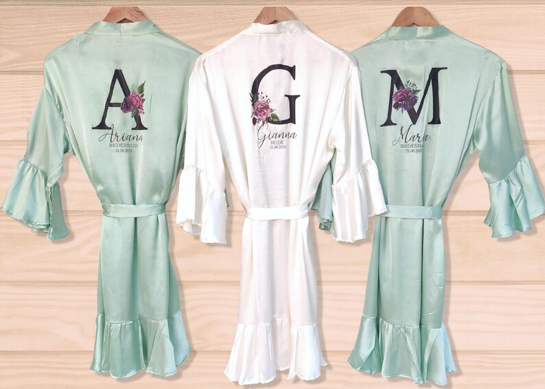 May include: Three satin robes with ruffled hems and bell sleeves. The robes are personalized with a large initial and the name of the wearer. The robes are in mint green and white.