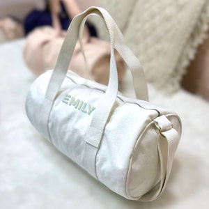 May include: White canvas duffel bag with a zipper closure and a shoulder strap. The bag has the name "EMILY" embroidered on the front in light blue.