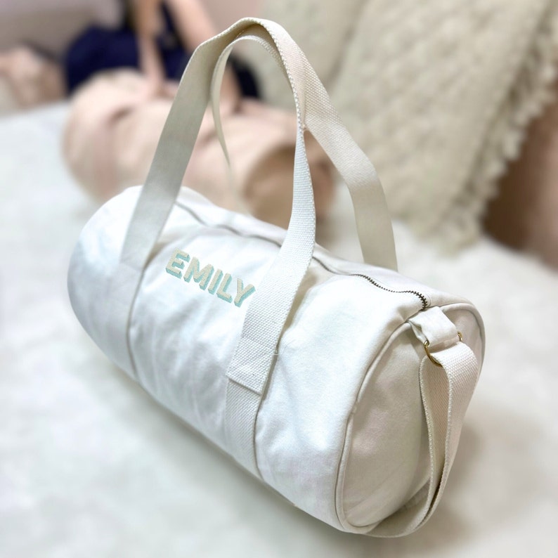 May include: White canvas duffel bag with a zipper closure and a shoulder strap. The bag has the name "EMILY" embroidered on the front in light blue.
