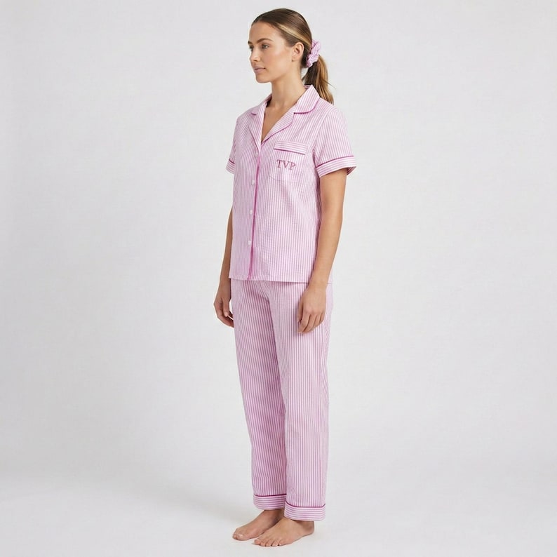 May include: A pink and white striped pajama set. The short-sleeved top has a collared neckline and the initials "TVP" embroidered on the chest. The pajama pants are long and have a matching stripe pattern. The set is trimmed with a darker pink piping.